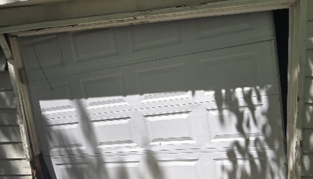 A broken white garage door off its tracks and hanging crookedly in a damaged frame.
