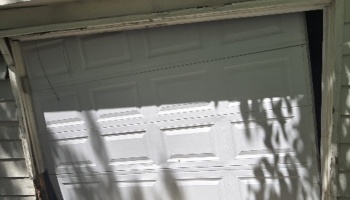 A broken white garage door off its tracks and hanging crookedly in a damaged frame.