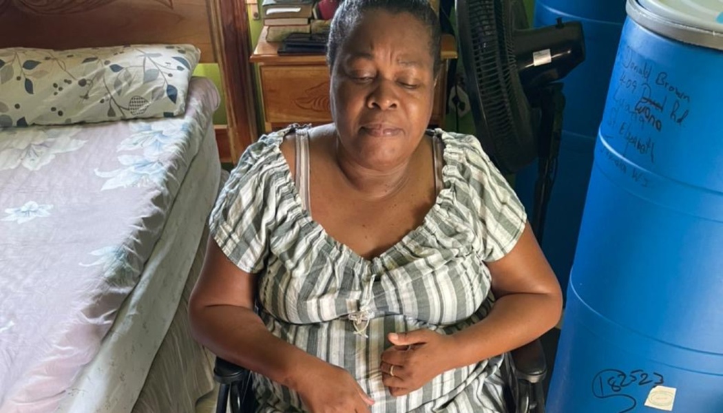 A woman sits in a wheelchair with her eyes closed inside a bedroom next to large blue storage barrels.