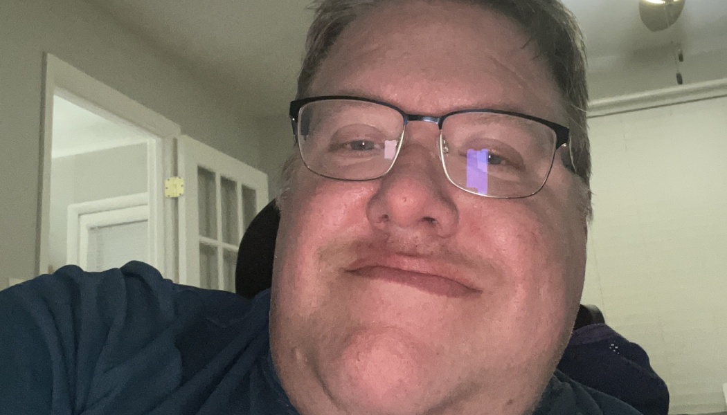 Close-up selfie of a smiling man wearing dark-framed glasses and a blue shirt.