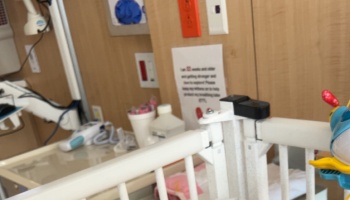 White crib railing in a hospital room with medical equipment and an informational sign posted on the wall.