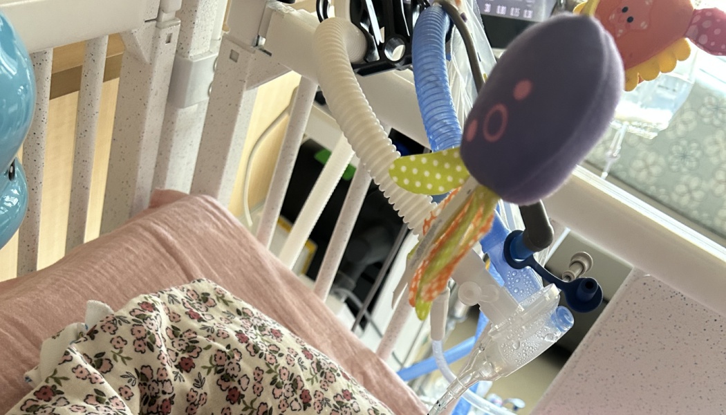 Medical breathing tubes and plush toys attached to a hospital crib with a floral blanket.