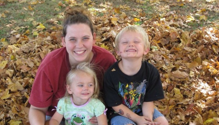 A smiling mother and her two young children sitting happily together in a pile of fallen autumn leaves.