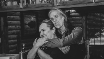 Black and white portrait of a long-haired man with a beard embracing a smiling woman from behind.