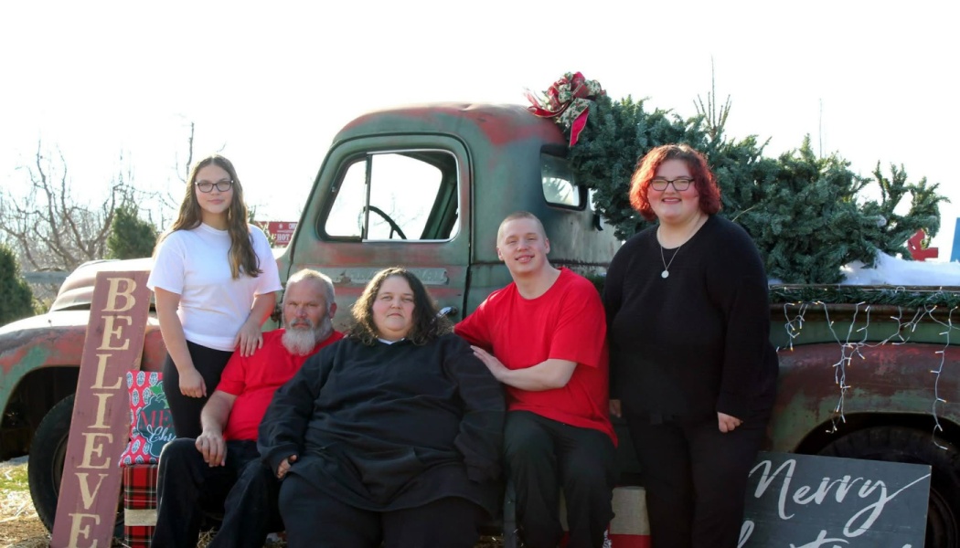 A family of five poses outdoors in front of a vintage green truck loaded with a Christmas tree.