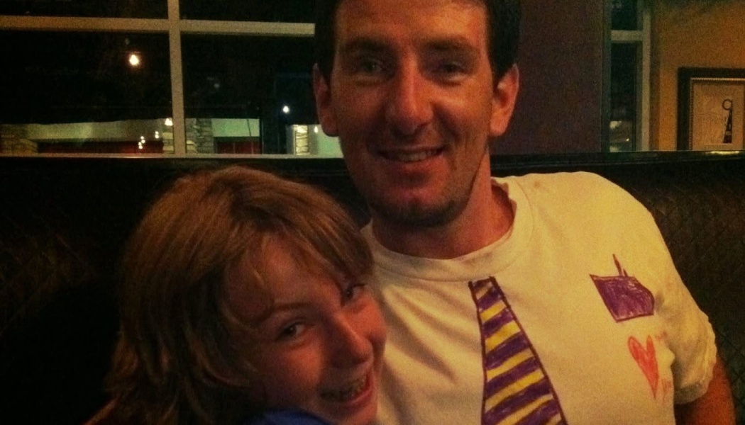 A smiling man and child lean together; the man wears a t-shirt featuring hand-drawn tie and heart designs.