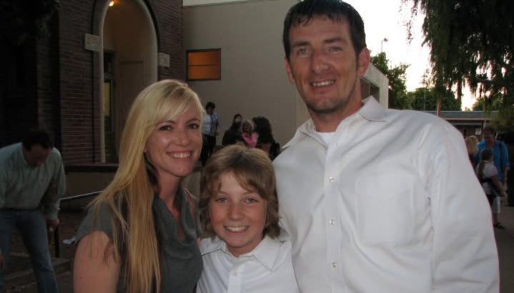 A smiling blonde woman, a young boy, and a man standing together outdoors in front of a building.