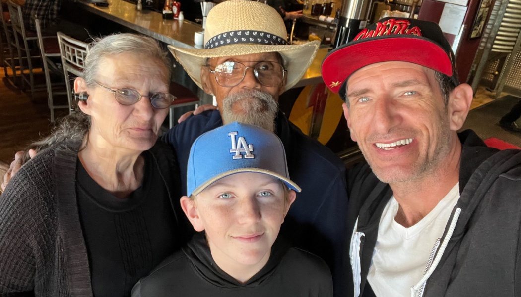 A smiling family of four, including two older adults and a boy in a cap, posing for a selfie indoors.