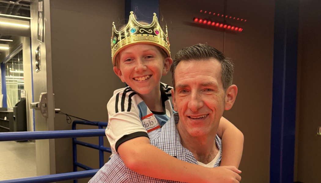 A smiling boy wearing a gold toy crown gives a piggyback hug to a smiling man.