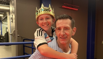 A smiling boy wearing a gold toy crown gives a piggyback hug to a smiling man.