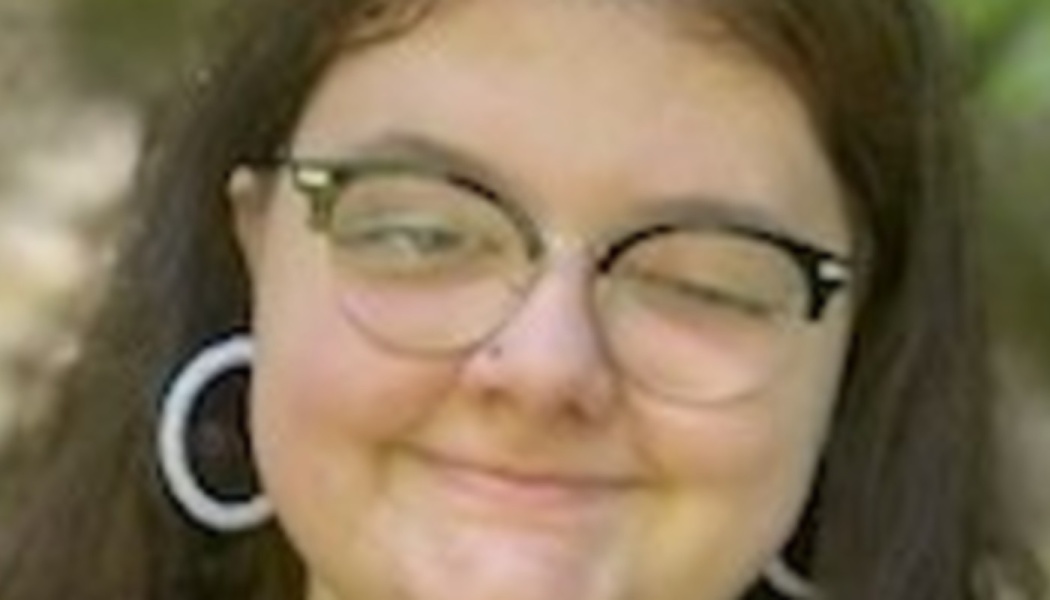 Close-up portrait of a smiling young person with glasses, long dark hair, and large hoop earrings.