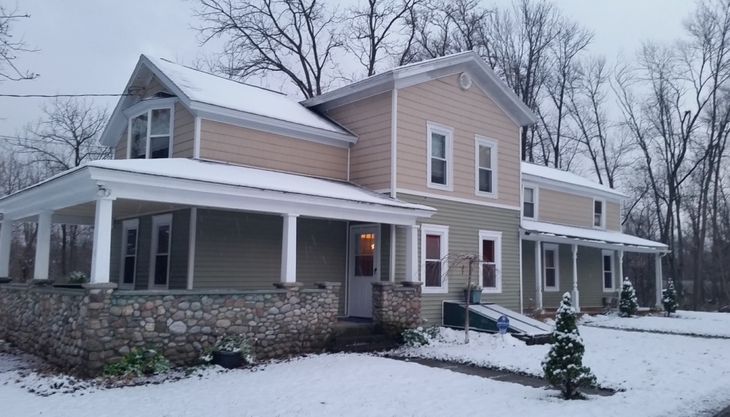 A snow-covered two-story house with green and beige siding and a stone porch on a winter day.