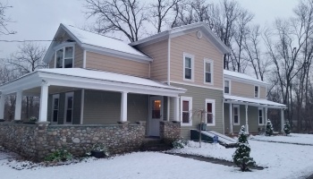 A snow-covered two-story house with green and beige siding and a stone porch on a winter day.