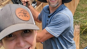 A smiling woman in a Limestone Farm cap takes a selfie with a man building a wooden structure outdoors.