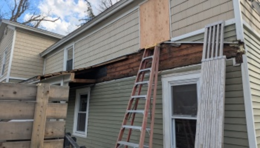 Damaged exterior wall of a house with a ladder leaning up to a boarded-up second-story doorway.