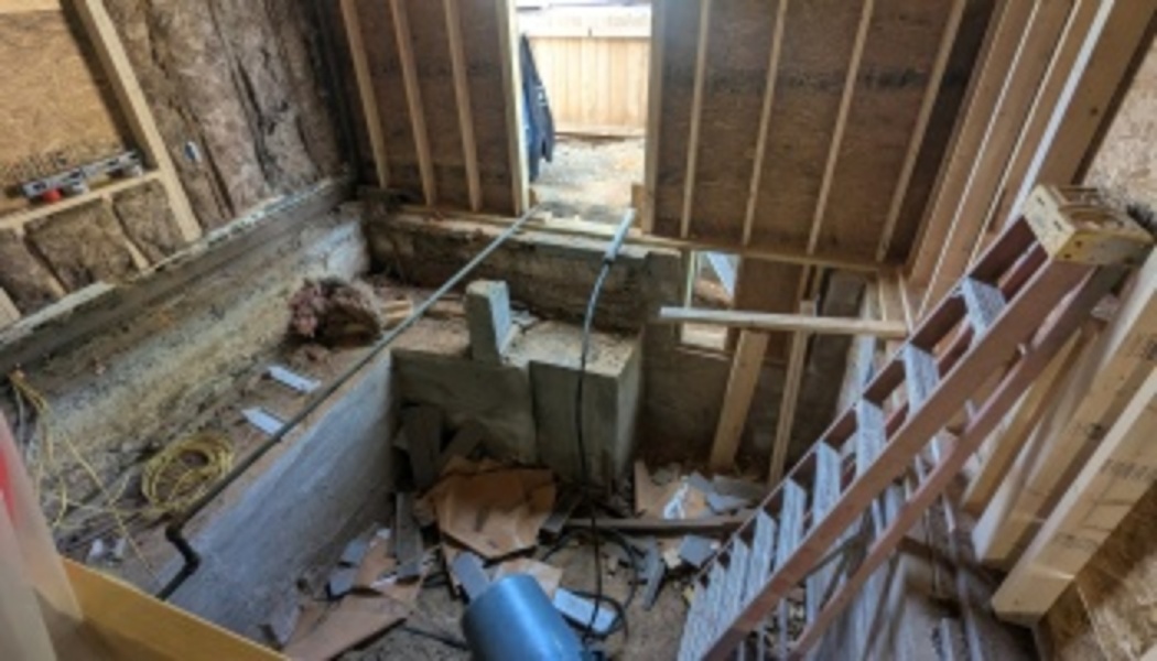 Exposed wooden wall studs, insulation, concrete foundation, and a wooden ladder inside a room under construction.