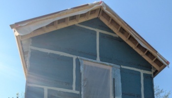 Exterior of a house under construction with blue weather wrap and exposed wooden roof overhangs against a blue sky.