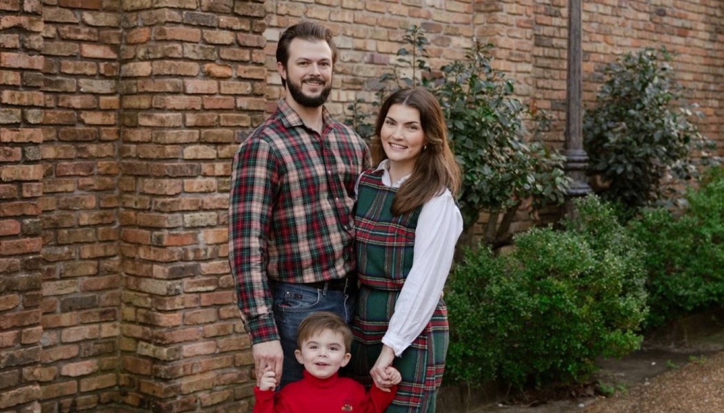 A smiling mother, father, and young son wearing matching festive plaid outfits pose in front of a brick wall.