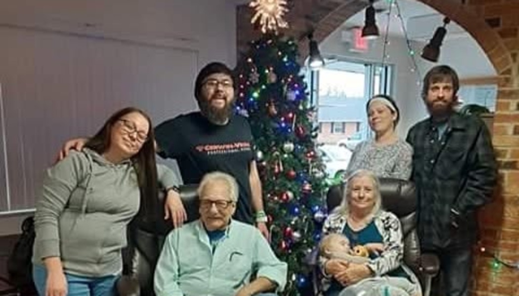 A multi-generational family smiles together while gathered around a lit, decorated Christmas tree.