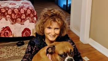 A smiling woman with styled brown hair sits on the floor behind her brown boxer dog.