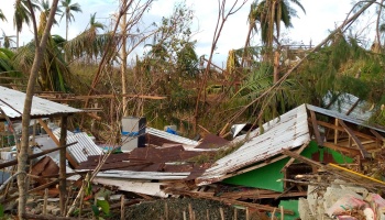 Destroyed homes with collapsed tin roofs, scattered debris, and broken trees after a severe tropical storm.