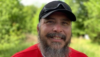 A smiling man with a beard, wearing a black cap and red shirt, with green foliage in the background.