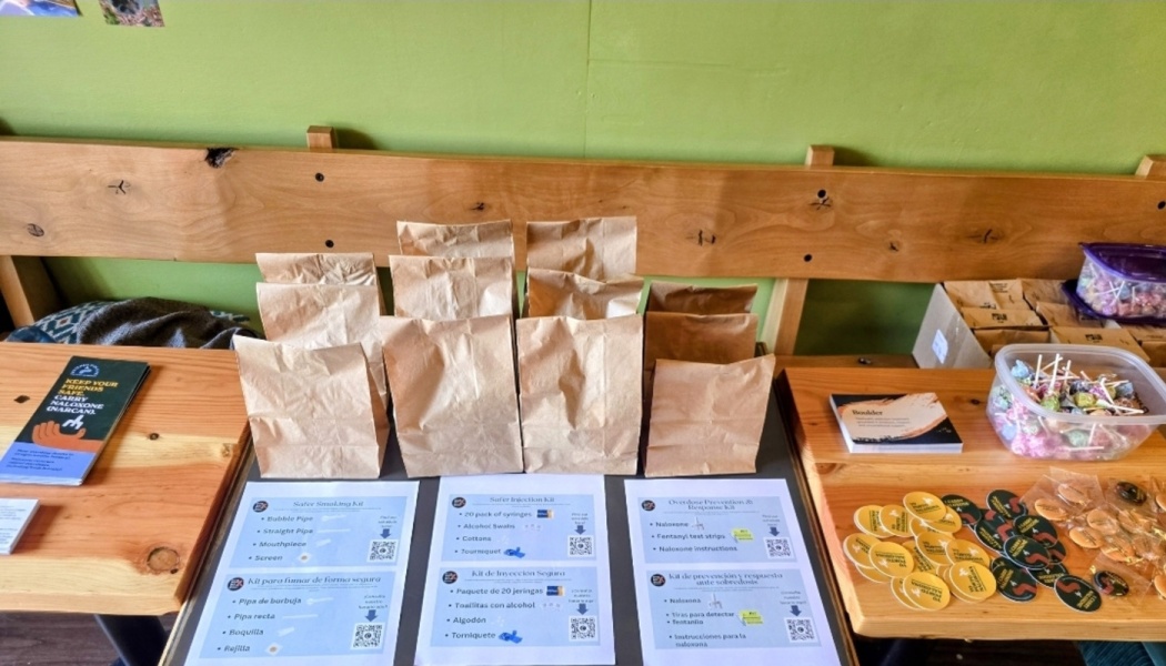 Brown paper bags with harm reduction kits lined up on a wooden table alongside informational flyers and buttons.