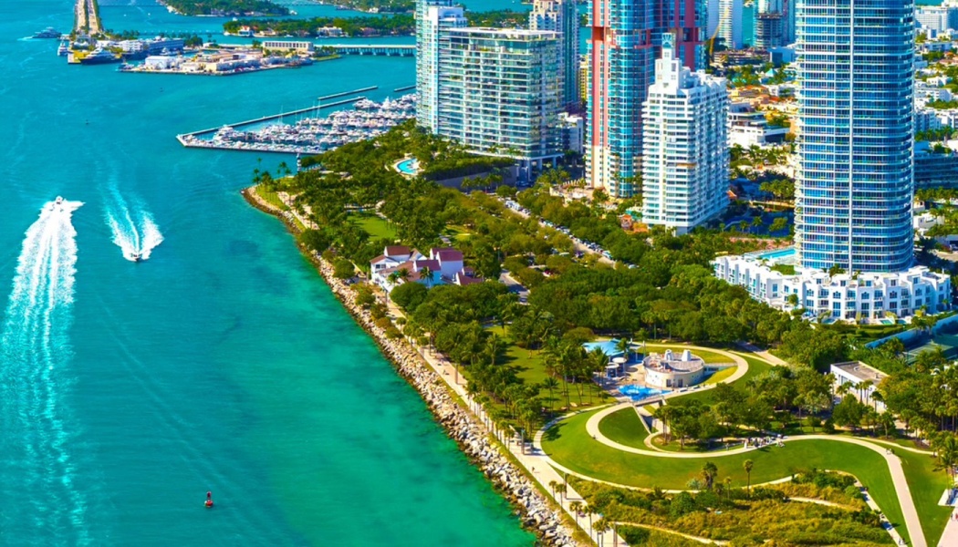 Aerial view of coastal skyscrapers, a green waterfront park, and speedboats in bright turquoise water.