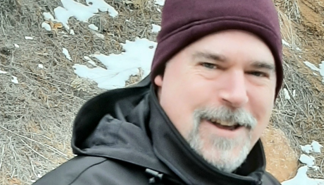 A smiling man with a gray goatee wearing a maroon beanie and black jacket outdoors near snowy ground.