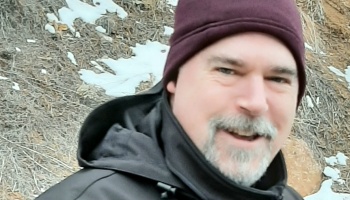 A smiling man with a gray goatee wearing a maroon beanie and black jacket outdoors near snowy ground.