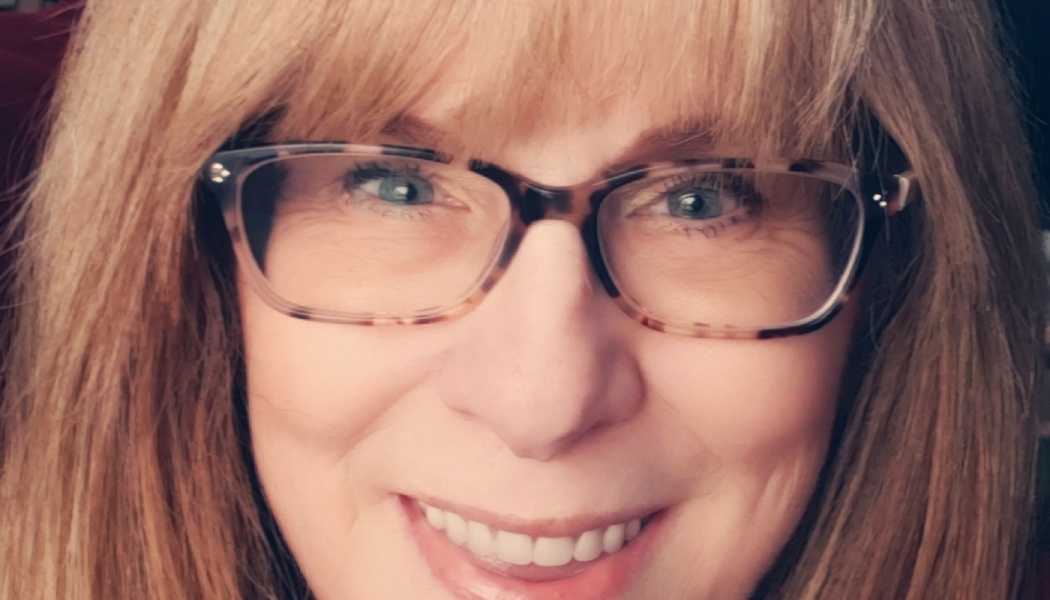 Close-up selfie of a smiling woman with blonde hair, bangs, and tortoiseshell glasses.