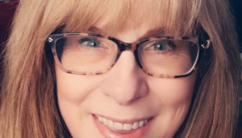 Close-up selfie of a smiling woman with blonde hair, bangs, and tortoiseshell glasses.