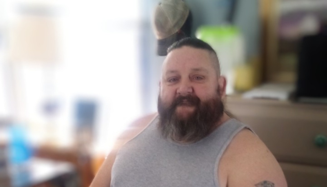 A man with a full beard and short dark hair wearing a grey tank top.