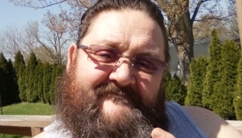 Close-up selfie of a smiling man with a full beard and glasses outdoors on a sunny day.