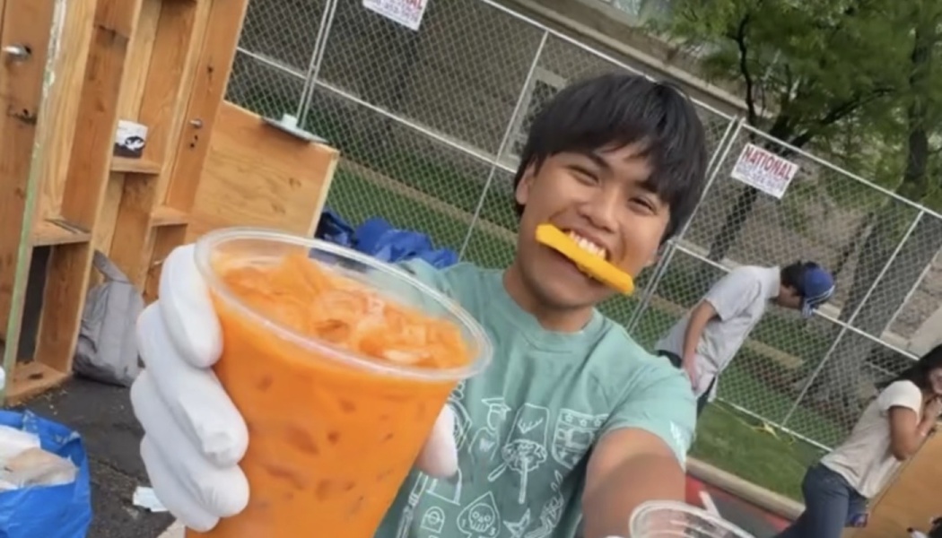Smiling volunteer holding out a cup of iced orange tea to the camera, with a snack playfully held in his mouth.