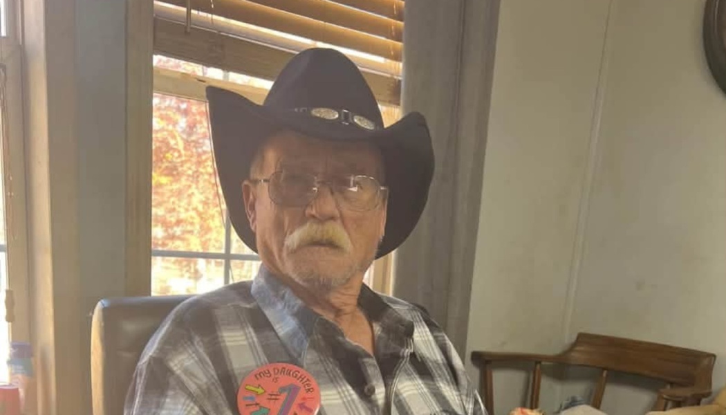 An older man wearing a black cowboy hat, glasses, and plaid shirt with a 'my daughter is #1' pin.