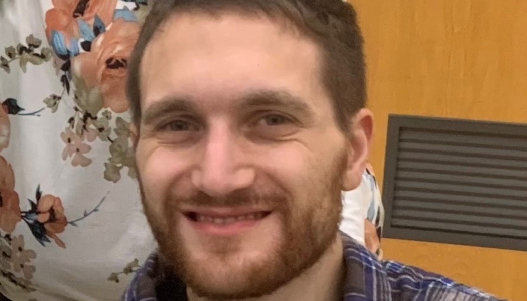 A smiling young man with short brown hair, a light beard, and a blue plaid shirt.
