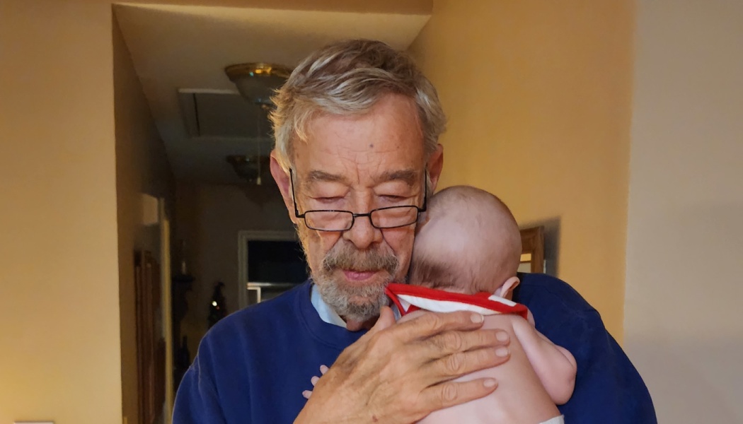 An elderly man with glasses tenderly embraces a baby resting on his shoulder.
