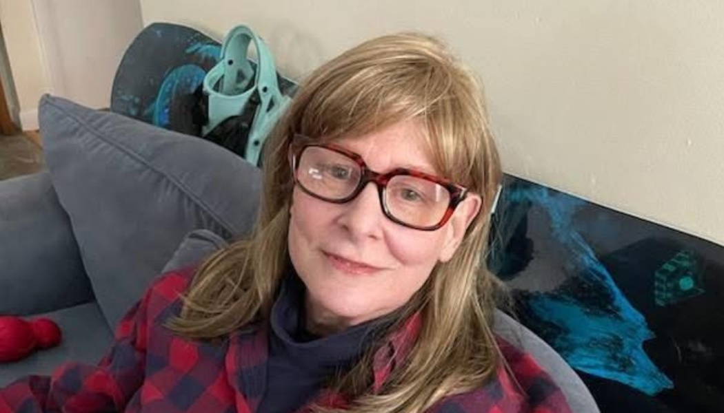 A smiling woman with blonde hair and glasses wearing a red plaid flannel shirt sitting on a couch.
