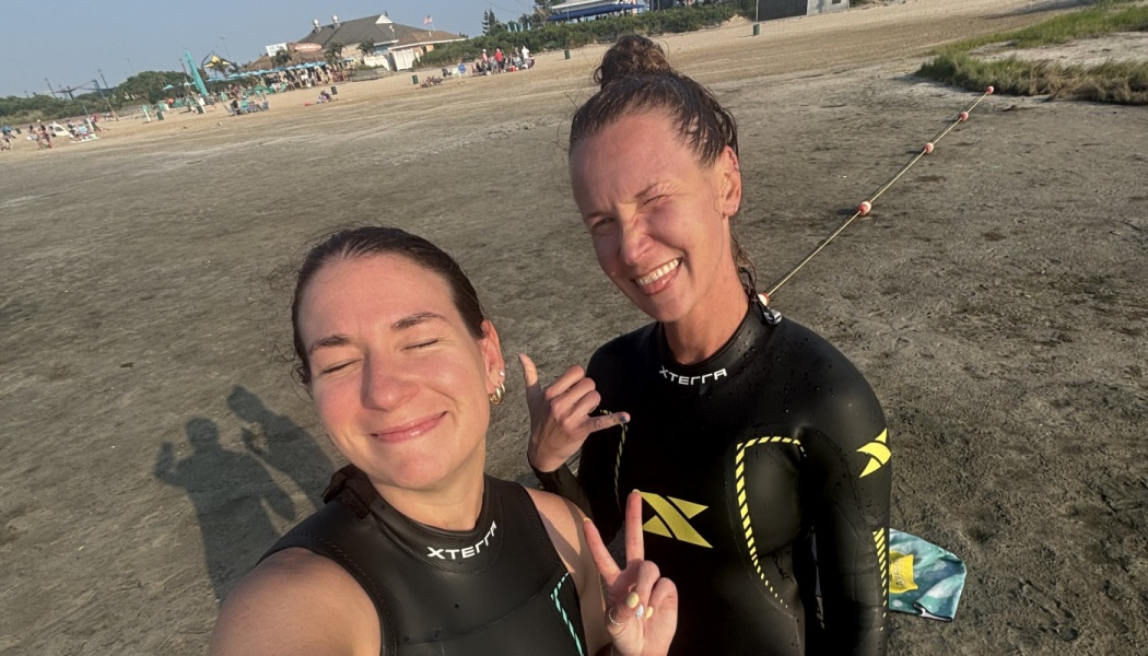 Two women in black wetsuits smile and make peace and shaka hand signs while taking a beach selfie.