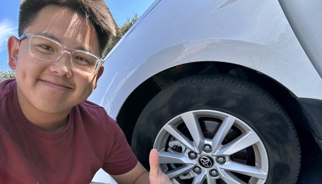 A smiling man wearing glasses gives a thumbs-up next to the tire of a white Toyota car.