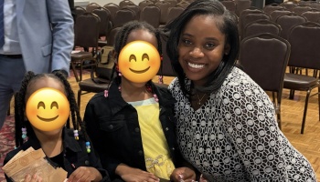 A smiling woman poses with two young girls with emoji-covered faces inside an event venue.