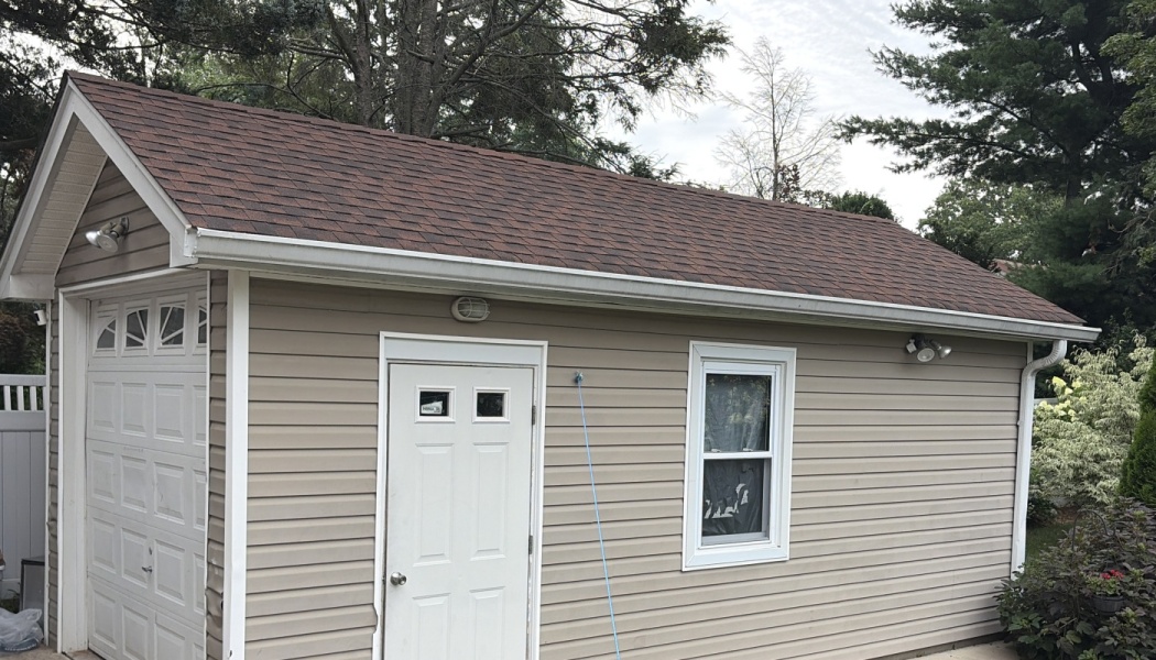 Tan vinyl-sided garage with a brown shingled roof, white side entrance door, single window, and white garage door.