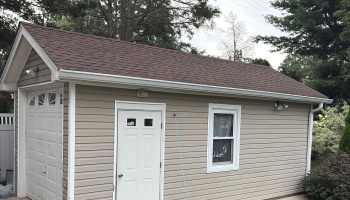 Tan vinyl-sided garage with a brown shingled roof, white side entrance door, single window, and white garage door.