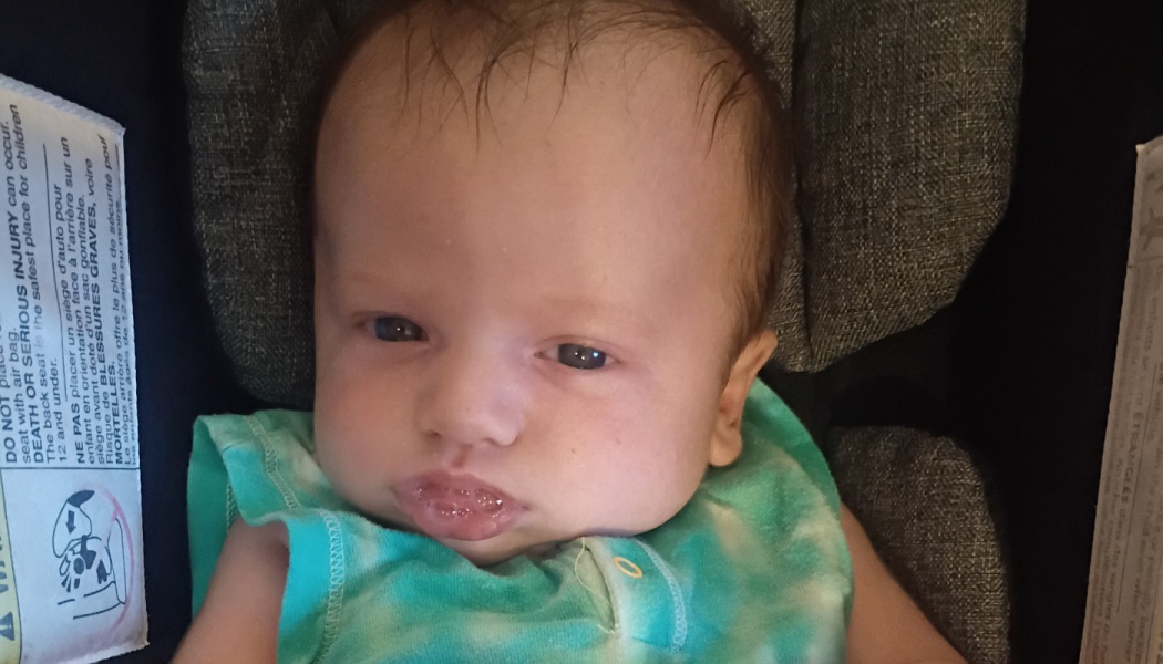 A newborn baby in a teal tie-dye top rests in a car seat and looks directly at the camera.