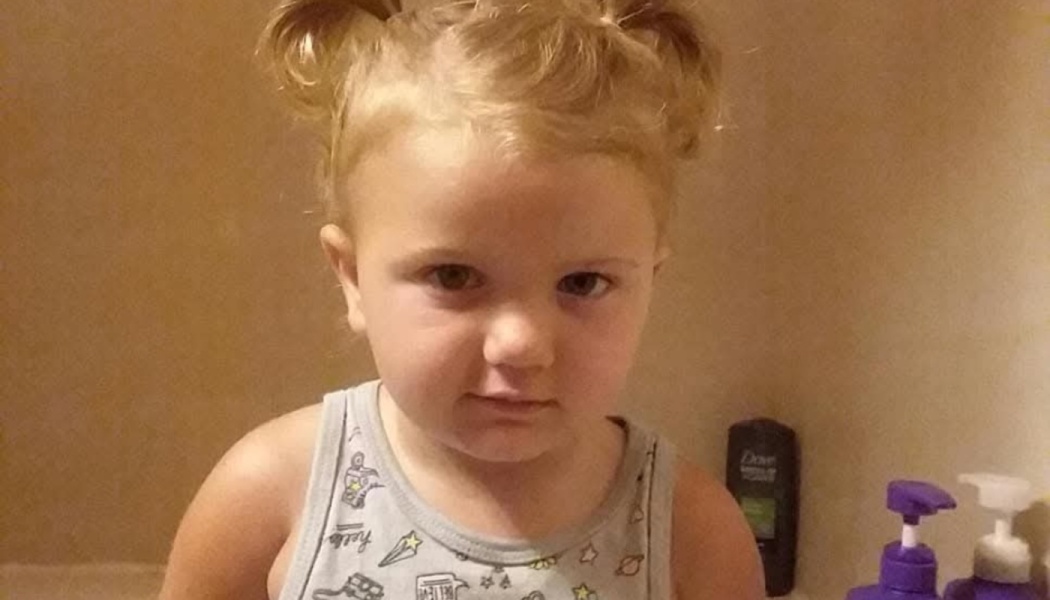 A blonde toddler with pigtails wearing a patterned tank top looks directly at the camera.
