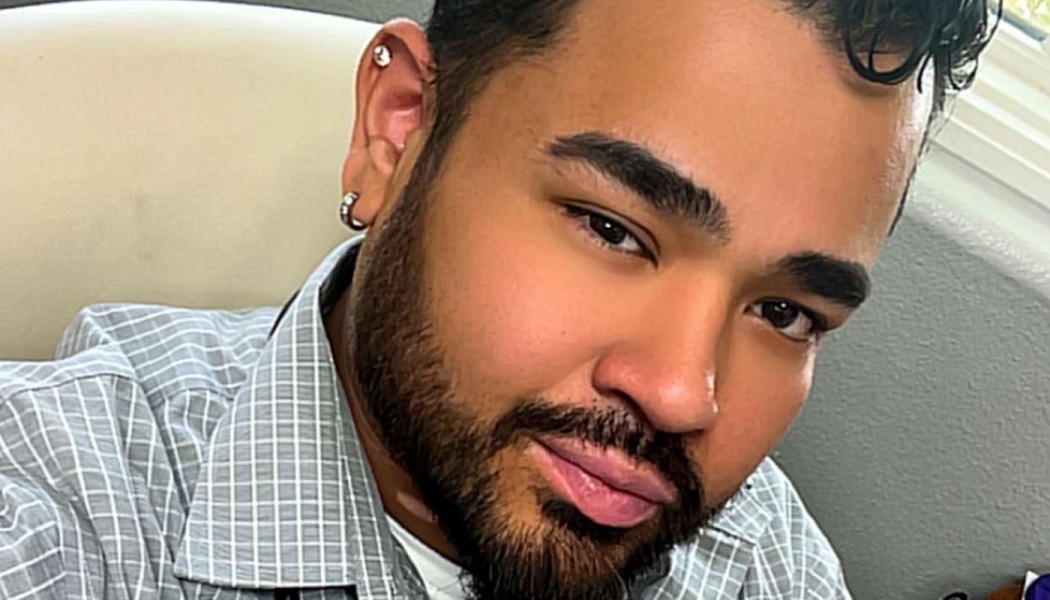 Close-up selfie of a man with a dark beard, ear piercings, wearing a grey plaid button-down shirt.