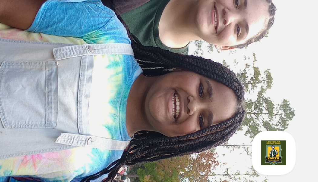 Two smiling youths outdoors, one wearing a colorful tie-dye shirt and overalls.