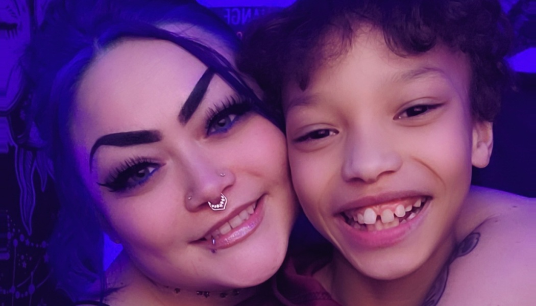 A smiling woman with facial piercings poses close to a happy young child under vibrant purple lighting.