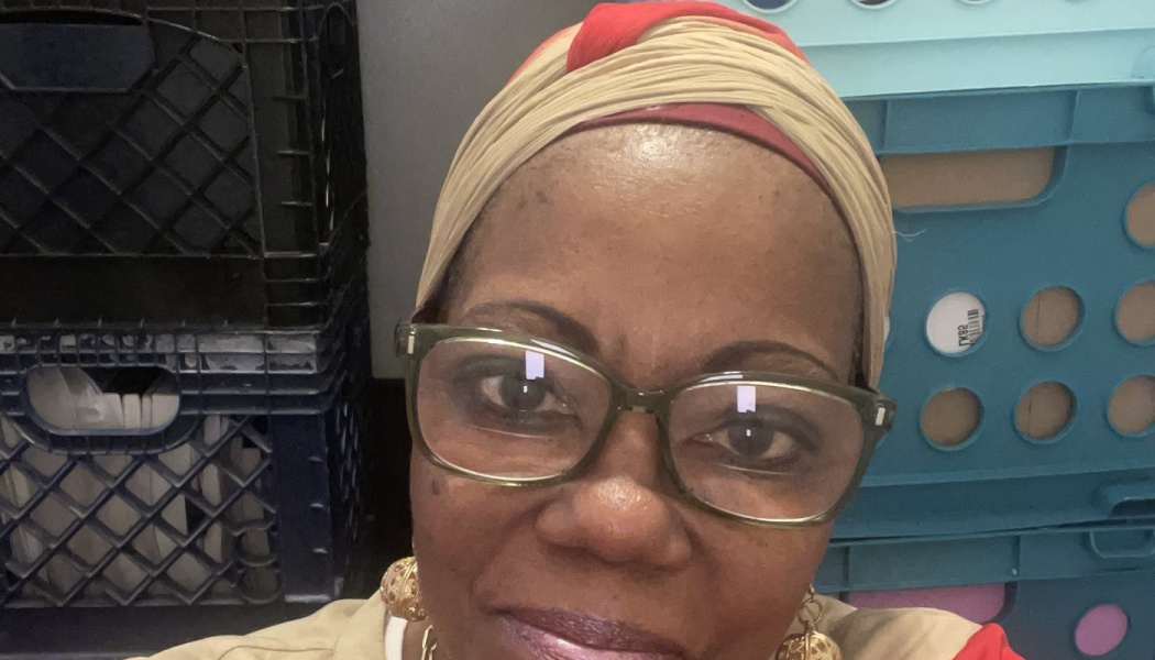 Close-up of a smiling Black woman wearing glasses and a beige headwrap with red accents.